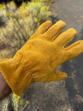 Load image into Gallery viewer, A close-up of a deerskin leather glove made sewn with the suede side out green gold leather, worn on a hand with an outdoor blurred background of brush and a path.
